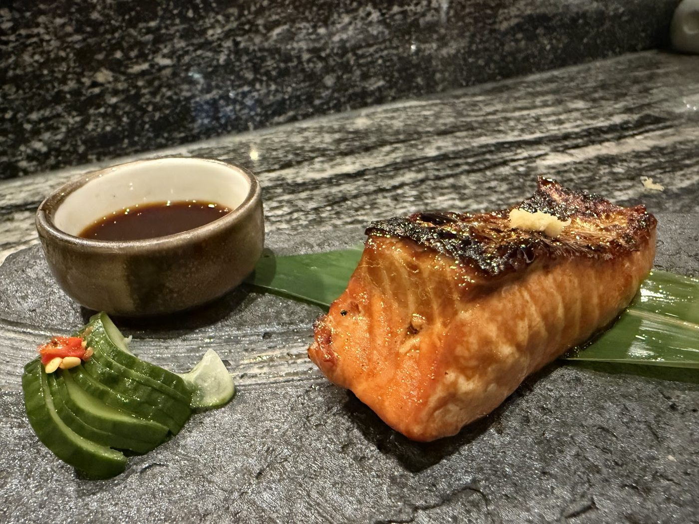 Grilled salmon fillet on a stone plate with dipping sauce and cucumber